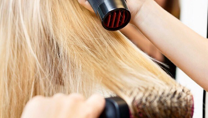 A hairstylist using a round brush and hairdryer to style a client's long blonde hair and apply makeup. - K. Charles & Co. in San Antonio and Schertz, TX