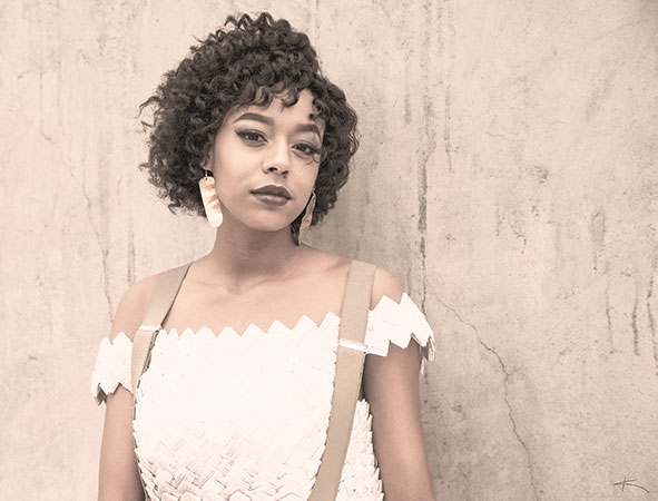 A young woman with curly hair, wearing a white dress and large earrings, stands before a textured beige wall. - K. Charles & Co. in San Antonio and Schertz, TX