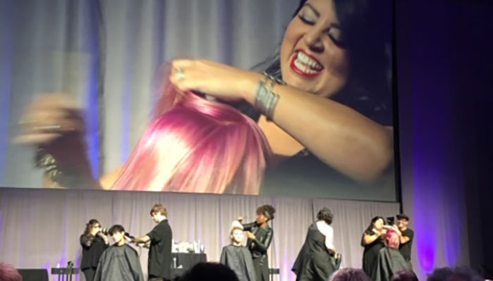 Hair stylists demonstrate hairstyling techniques on models at an event, with a large screen displaying a close-up of a smiling woman having her hair styled. - K. Charles & Co. in San Antonio and Schertz, TX