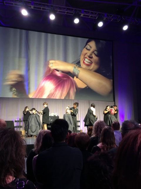 A hairstyling demonstration on stage at a convention, with a large screen displaying a smiling woman holding colorful hair extensions. audience members watch attentively. - K. Charles & Co. in San Antonio and Schertz, TX