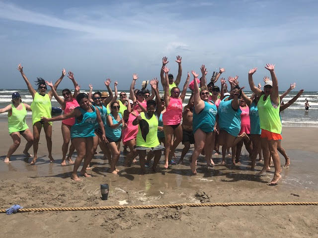 A group of people with styled hair in colorful beachwear cheerfully posing with raised arms on a sandy beach, with waves in the background. - K. Charles & Co. in San Antonio and Schertz, TX