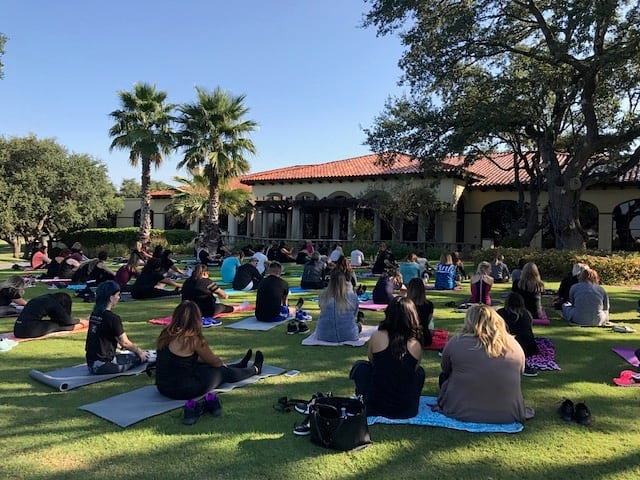 Outdoor yoga class with participants seated on mats in a park, facing an instructor, with trees and a building in the background. - K. Charles & Co. in San Antonio and Schertz, TX