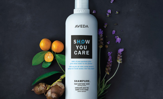 Aveda shampoo bottle with the text "show you care" surrounded by orange fruits, a leaf, and scattered purple flowers on a dark background, perfect for any salon. - K. Charles & Co. in San Antonio and Schertz, TX