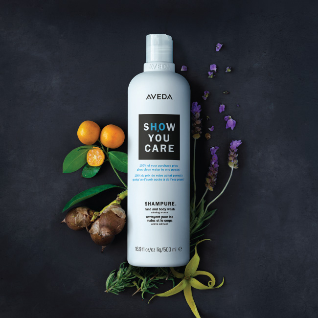 Aveda shampoo bottle with the text "show you care" surrounded by purple flowers, citrus fruits, and green leaves on a dark background. - K. Charles & Co. in San Antonio and Schertz, TX