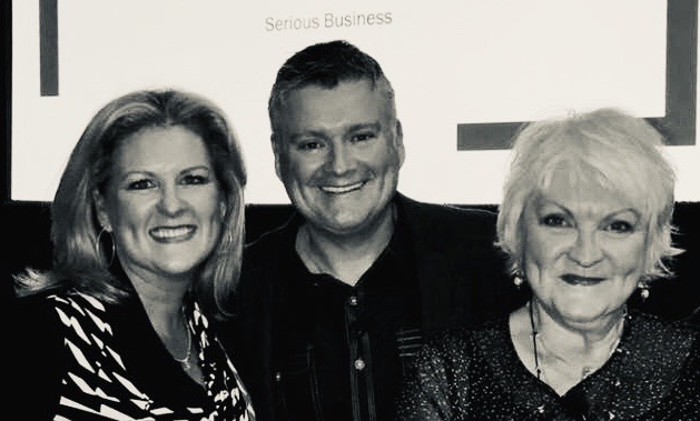 Three smiling adults, two women and one man, pose for a photo in black and white; a screen behind them reads "serious hairstyle. - K. Charles & Co. in San Antonio and Schertz, TX