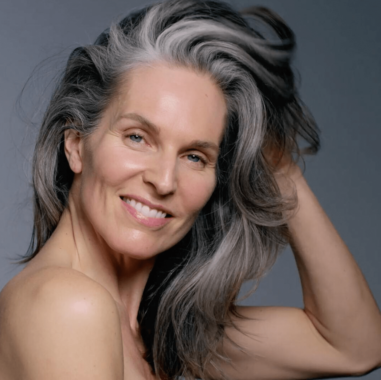 Portrait of a smiling middle-aged woman with a stylish salon hairstyle, featuring long, flowing gray hair against a gray background. - K. Charles & Co. in San Antonio and Schertz, TX