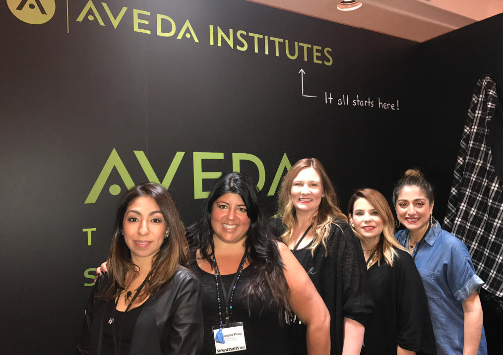 Five women smiling at a conference booth with the aveda institute logo and the slogan "it all starts here!" in the background. - K. Charles & Co. in San Antonio and Schertz, TX