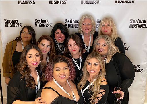 Group of ten smiling women posing for a photo at a "serious business" conference, standing in front of a branded backdrop. - K. Charles & Co. in San Antonio and Schertz, TX