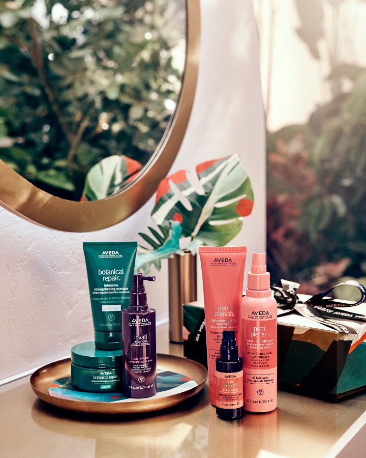 A collection of aveda beauty products arranged on a tray before a round mirror, with lush greenery in the background. - K. Charles & Co. in San Antonio and Schertz, TX