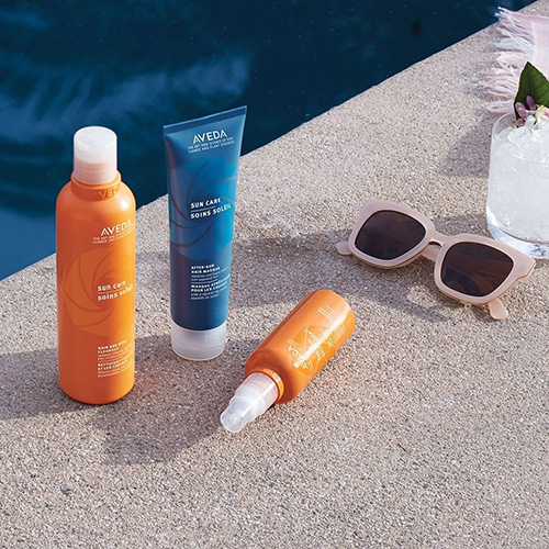 Three Aveda sun care products arranged beside sunglasses and a flower by a swimming pool, ideal for maintaining hair color protection in the sun. - K. Charles & Co. in San Antonio and Schertz, TX