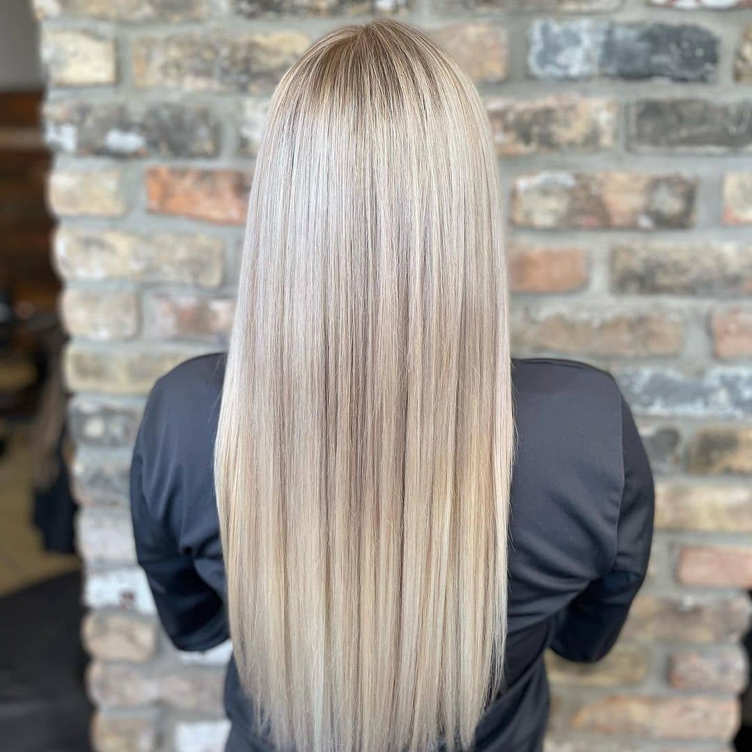 A person with long, straight blonde hair colored at a salon, standing in front of a stone wall, viewed from behind. - K. Charles & Co. in San Antonio and Schertz, TX