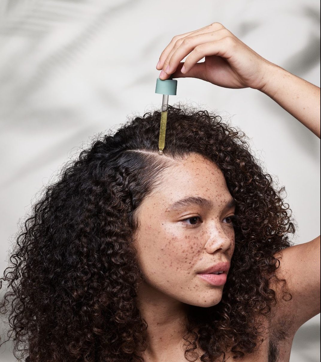 A woman with curly hair applies oil to her scalp using a dropper. - K. Charles & Co. in San Antonio and Schertz, TX