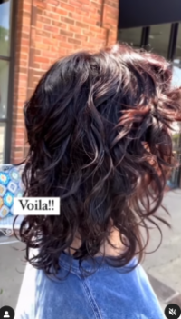 Back view of a person with a salon-styled shoulder-length curly hairstyle highlighted with red tones, standing outside a brick building, with a caption that reads "voila!!". - K. Charles & Co. in San Antonio and Schertz, TX