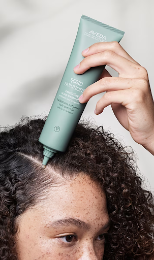 A person applying aveda scalp solutions serum to their scalp from a green tube, focusing on a section of curly hair. - K. Charles & Co. in San Antonio and Schertz, TX