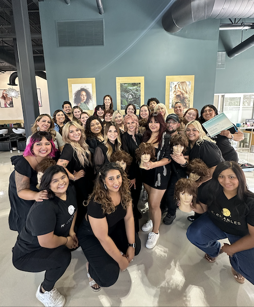 Group of hairstylists in black uniforms posing with mannequin heads at a salon, smiling at the camera. - K. Charles & Co. in San Antonio and Schertz, TX