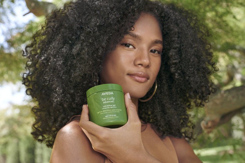 A young woman with curly hair holding a jar of aveda be curly hair product, standing outdoors with greenery in the background. - K. Charles & Co. in San Antonio and Schertz, TX