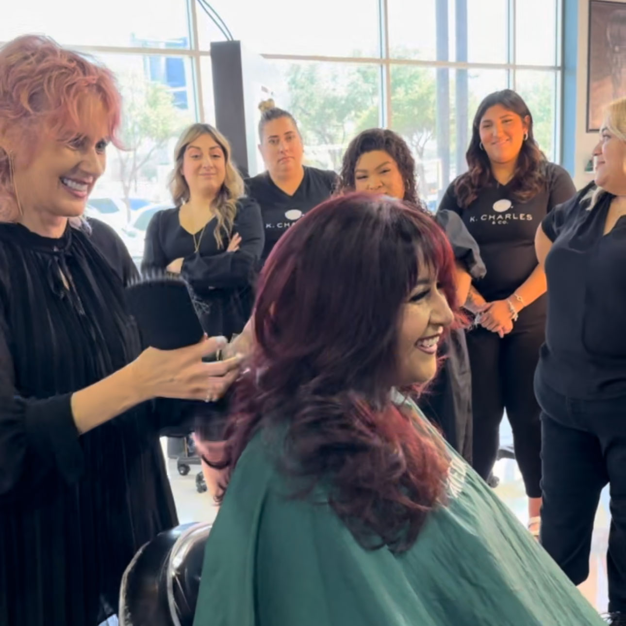A group of hairstylists watch as a woman with red hair gets her hair styled by another smiling hairstylist in a salon. - K. Charles & Co. in San Antonio and Schertz, TX
