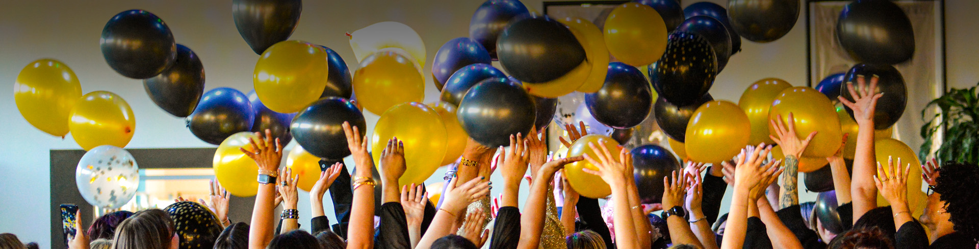 A crowd of people raise their hands towards black and gold balloons floating in the air at an indoor event. - K. Charles & Co. in San Antonio and Schertz, TX