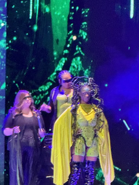 Three individuals stand on stage; one in a dramatic yellow outfit with elaborate headpiece, and two others in dark clothing, with a green and black abstract background behind them. - K. Charles & Co. in San Antonio and Schertz, TX