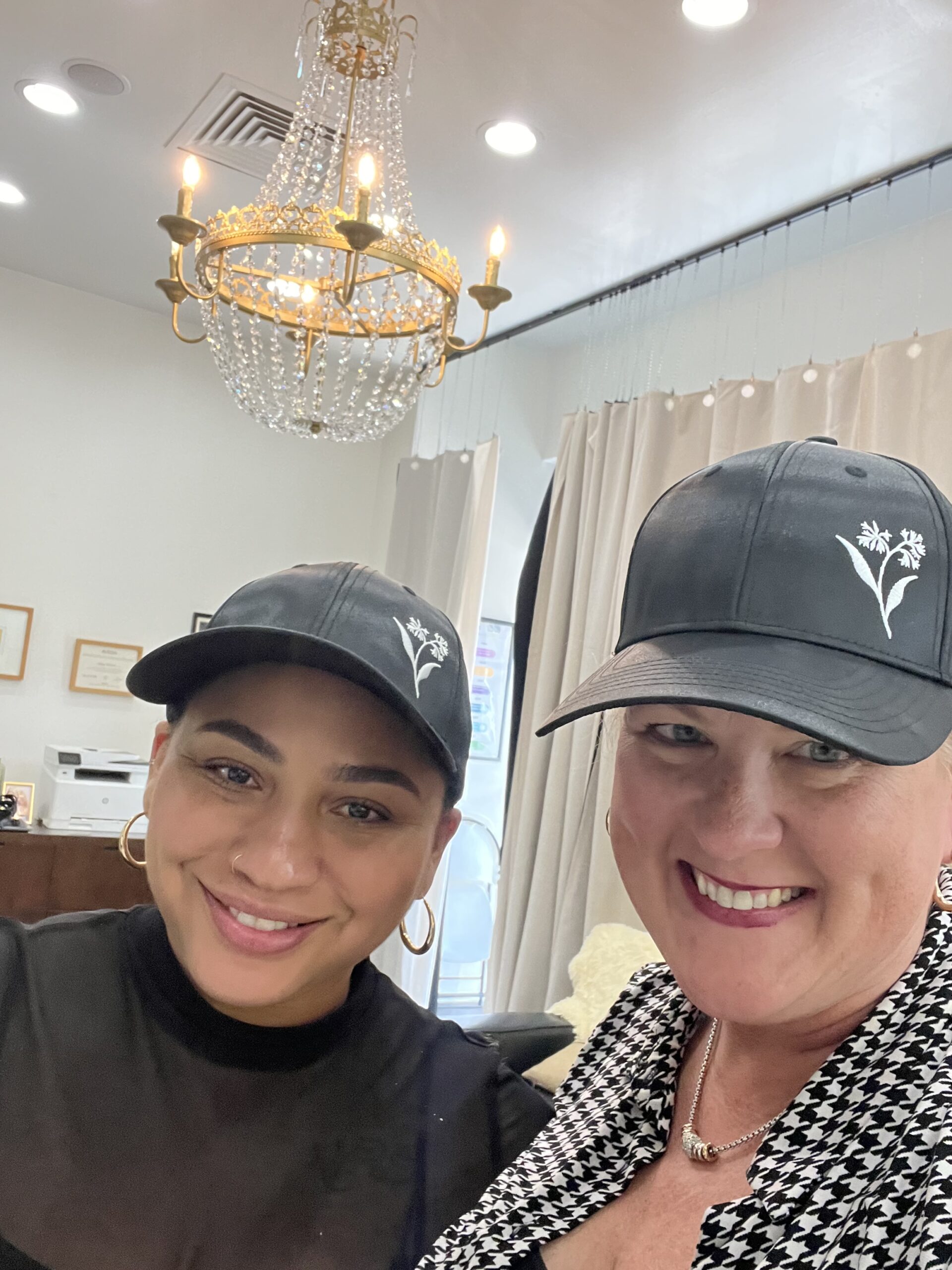 Two women wearing black hats with a white floral logo smile indoors in a well-lit room with a chandelier and framed certificates on the wall. - K. Charles & Co. in San Antonio and Schertz, TX
