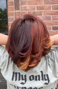 A person with shoulder-length, layered hair dyed black, red, and orange stands facing a brick wall, arms raised behind their head. The back of their shirt reads "My only". - K. Charles & Co. in San Antonio and Schertz, TX
