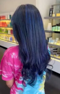 Person with long, wavy, dark blue hair stands in a store, wearing a pink and blue tie-dye shirt. Shelves with colorful bottles line the background. - K. Charles & Co. in San Antonio and Schertz, TX