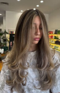 A person with long, layered, wavy ash-blonde hair is standing in a brightly lit salon, wearing a light-colored top. - K. Charles & Co. in San Antonio and Schertz, TX