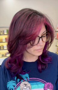 Person with shoulder-length, layered magenta and purple hair, wearing glasses and a blue T-shirt, looks downward in a brightly lit room. - K. Charles & Co. in San Antonio and Schertz, TX