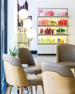A salon waiting area with beige chairs, a small table, three globe floor lamps, and shelves displaying assorted hair products arranged by color. - K. Charles & Co. in San Antonio and Schertz, TX