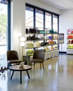 A modern salon waiting area with two brown chairs, a round wooden table, and shelves displaying various hair products by large windows. - K. Charles & Co. in San Antonio and Schertz, TX