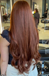 Person with long, wavy auburn hair stands in a modern salon, facing away from the camera. Salon chairs, mirrors, and another person are visible in the background. - K. Charles & Co. in San Antonio and Schertz, TX