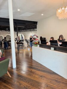 Reception desk with two women working at computers in a modern salon; clients and stylists are visible in the background. - K. Charles & Co. in San Antonio, TX