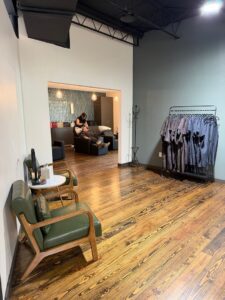 Modern salon interior with wooden floors, green chairs, a rack of hanging capes, and a stylist washing a client’s hair in the background. Warm lighting and minimalist decor. - K. Charles & Co. in San Antonio, TX