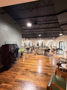 Interior view of a modern hair salon with wooden floors, styling chairs, large mirrors, and a rack of capes on the left. - K. Charles & Co. in San Antonio, TX