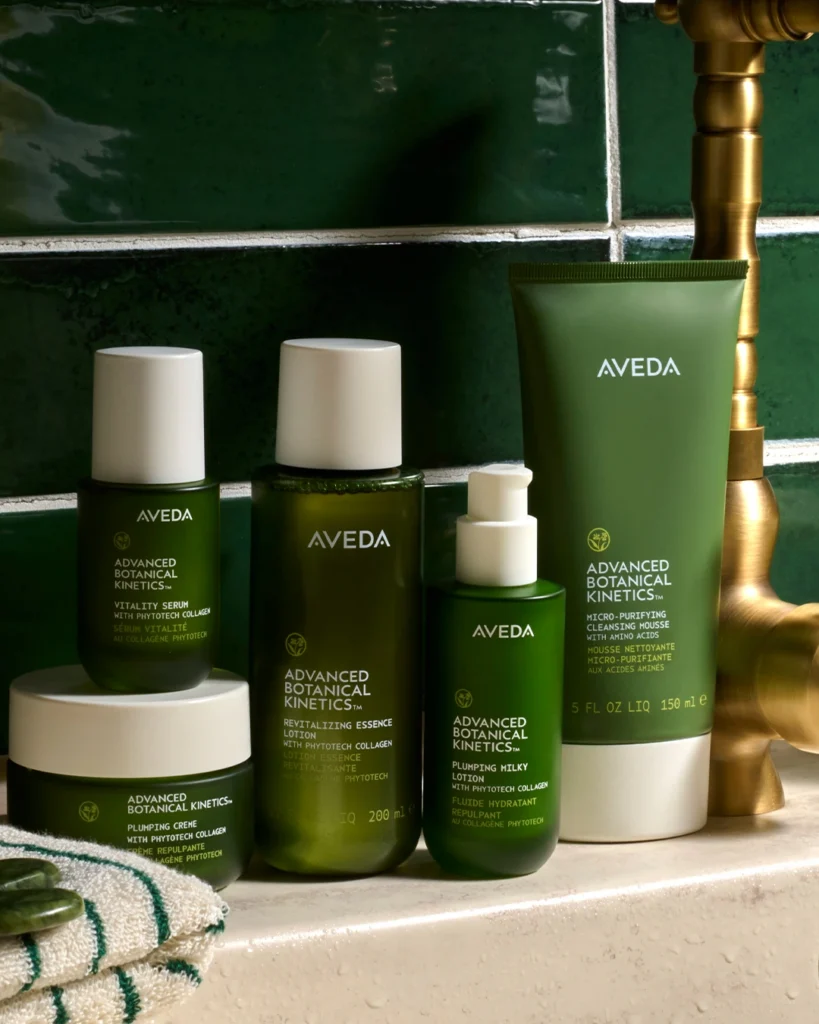 Five green Aveda Advanced Botanical Kinetics skincare products are arranged on a tiled bathroom counter next to a gold faucet, with a dark green tiled wall in the background. - K. Charles & Co. in San Antonio, TX